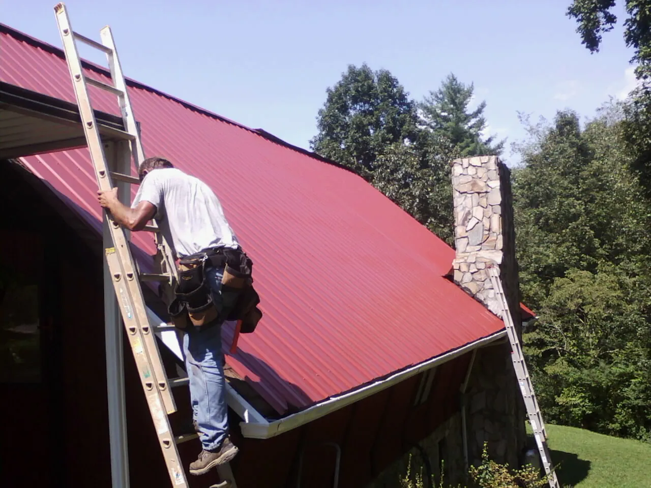 Roofer on a ladder beside a red metal roof and stone chimney — Soffit & Fascia Repair by Roofing Brothers in Staunton